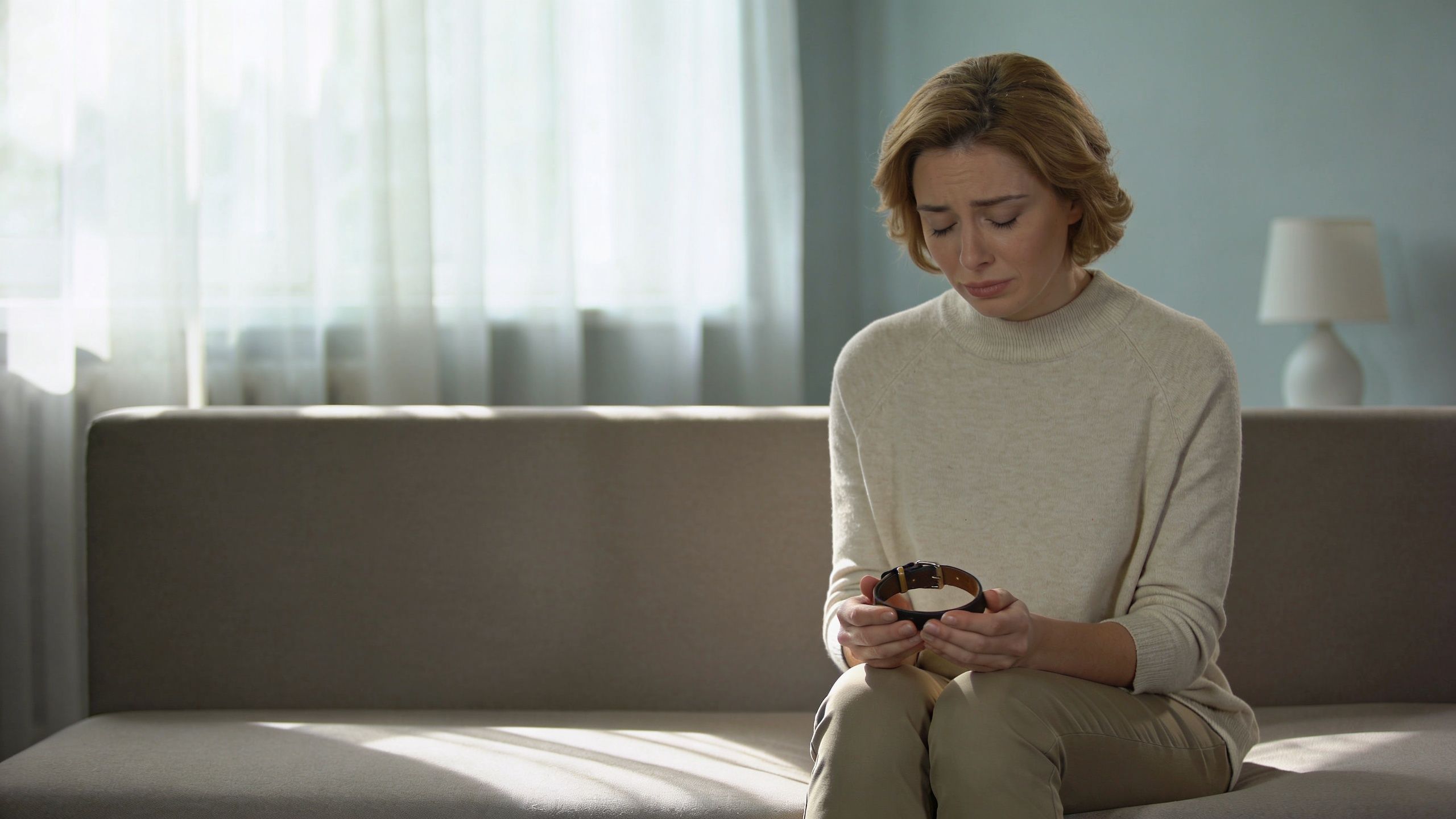 A sad woman sitting on a couch holding a dog collar, grieving the loss of her pet.